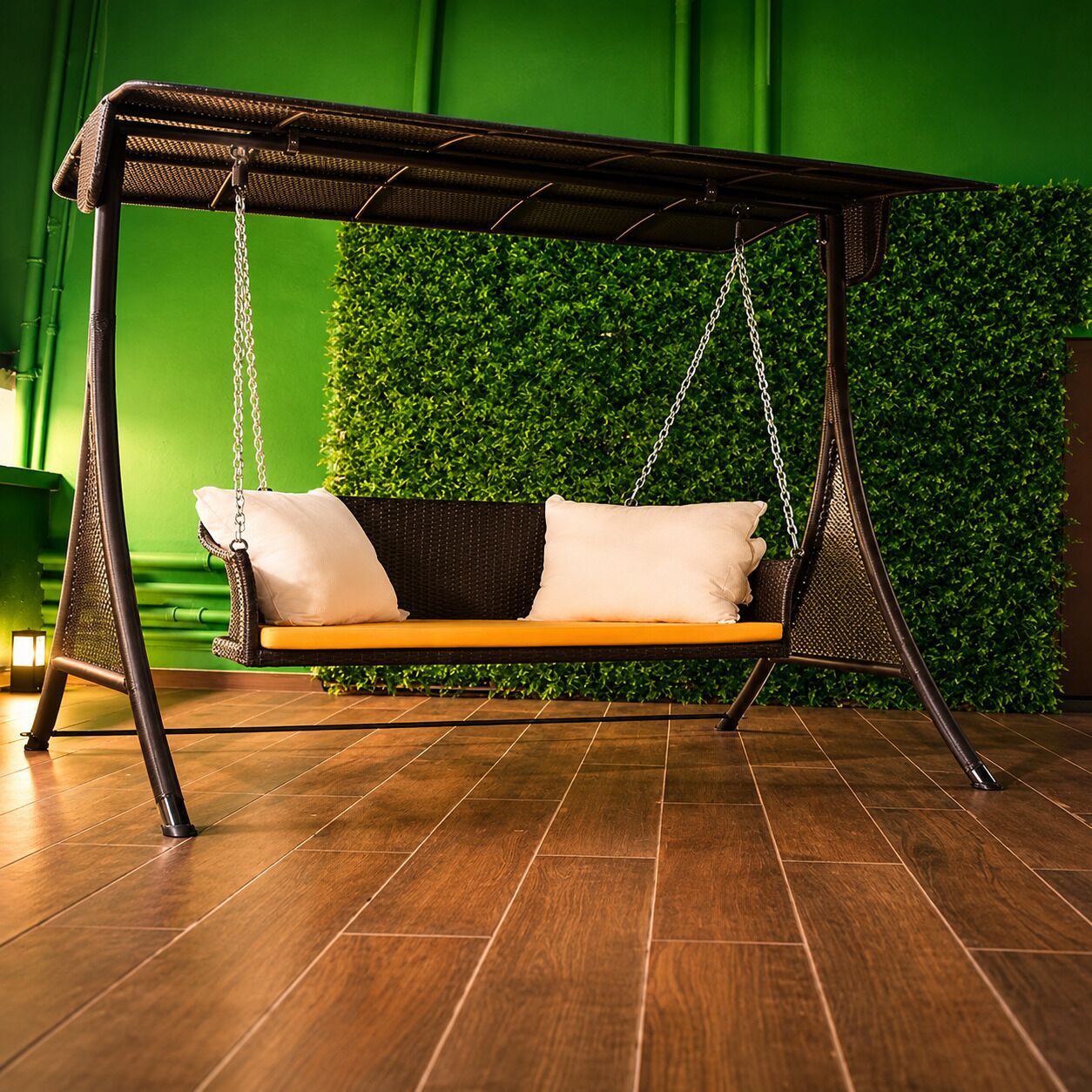 Silla colgante con asiento de madera y cojines, frente a una pared verde con seto.