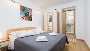A neatly made bed with a gray bedspread, a wooden headboard, and a nightstand with a lamp. There is a wardrobe with a mirror and a bathroom visible through an open door. - Downtown Split City Apartment (Split)