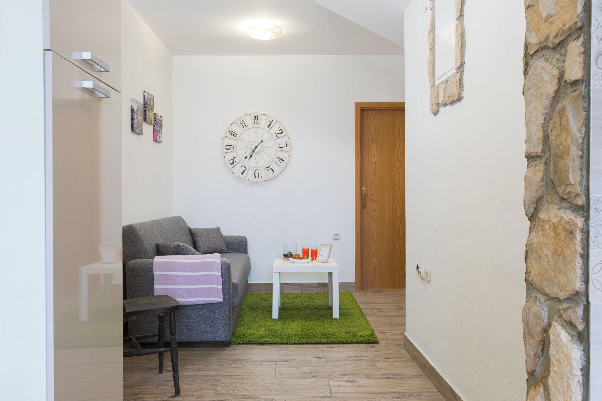 A small, modern living room with a grey sofa, a white coffee table, and a green rug. A large wall clock is visible, and a wooden door is in the background.