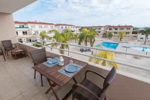 A balcony with a dining table set for two, overlooking a swimming pool and a residential area with palm trees. - Narcissos Nissi Beach Apartment D9 (Ayia Napa)