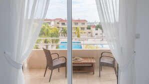 A balcony with two wicker chairs and a wooden table, overlooking a swimming pool and buildings. - Narcissos Nissi Beach Apartment D9 (Ayia Napa)