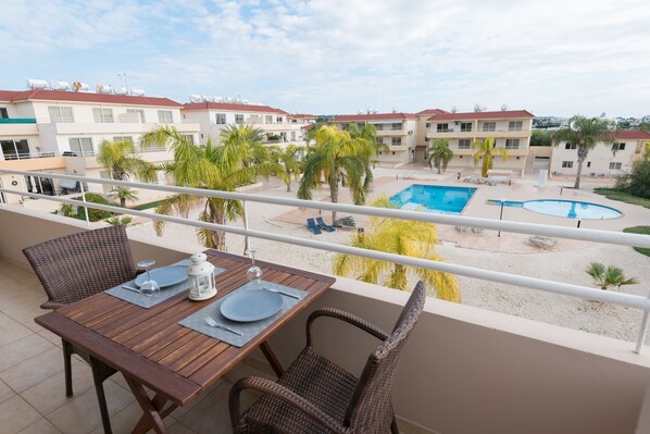 A balcony with a wooden table set for two, overlooking a swimming pool and a courtyard with palm trees. - Narcissos Nissi Beach Apartment D9 (Ayia Napa)