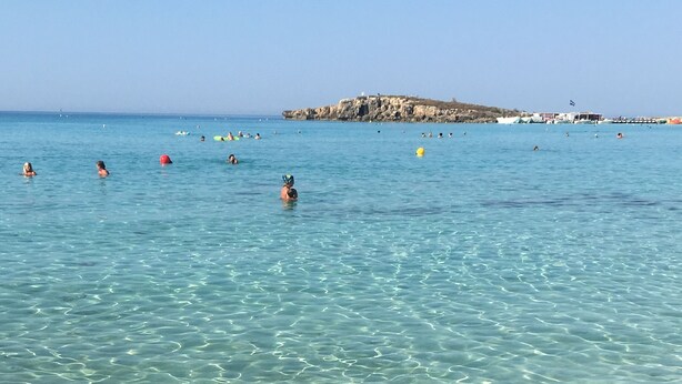 A clear beach with shallow water, a rocky outcrop in the distance, and boats on the water. - Narcissos Nissi Beach Apartment D9 (Ayia Napa)