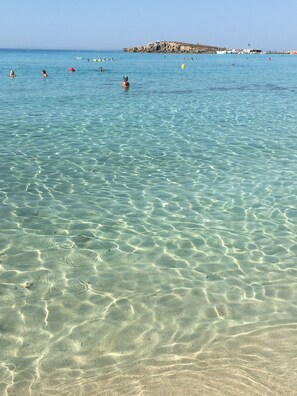 A clear beach with shallow water, a rocky outcrop in the distance, and boats on the water. - Narcissos Nissi Beach Apartment D9 (Ayia Napa)