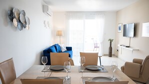 A dining area with a table set for four, a blue sofa, and a TV mounted on the wall. - Narcissos Nissi Beach Apartment D9 (Ayia Napa)