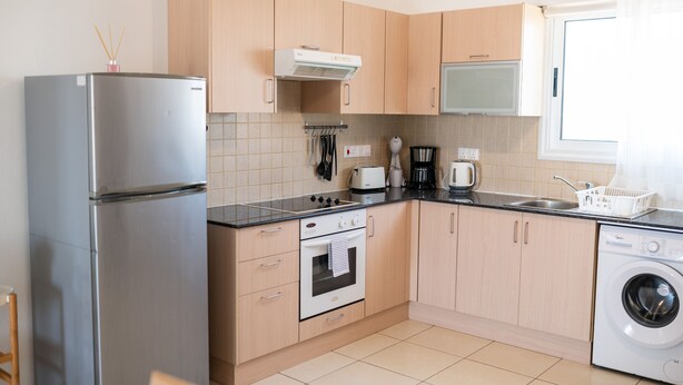 A modern kitchen with a stainless steel refrigerator, a built-in oven, a washing machine, and wooden cabinets. - Narcissos Nissi Beach Apartment D9 (Ayia Napa)