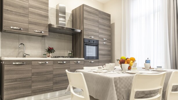 Une cuisine moderne avec des armoires en bois, une hotte de cuisine en acier inoxydable et un four encastré. On y trouve une table à manger avec des chaises et un panier de fruits posé sur la table.