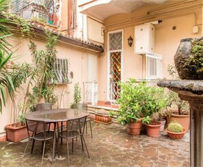 Une terrasse dallée de pierres, avec une table ronde et des chaises, entourée de plantes en pot et d’une petite fontaine.