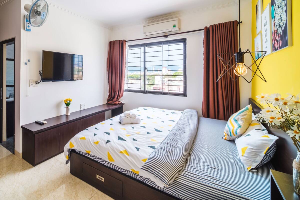 Une chambre d’hôtel comprenant un lit, une télévision, une fenêtre avec des rideaux, une table de chevet ornée d’un vase de fleurs et un climatiseur fixé au mur.