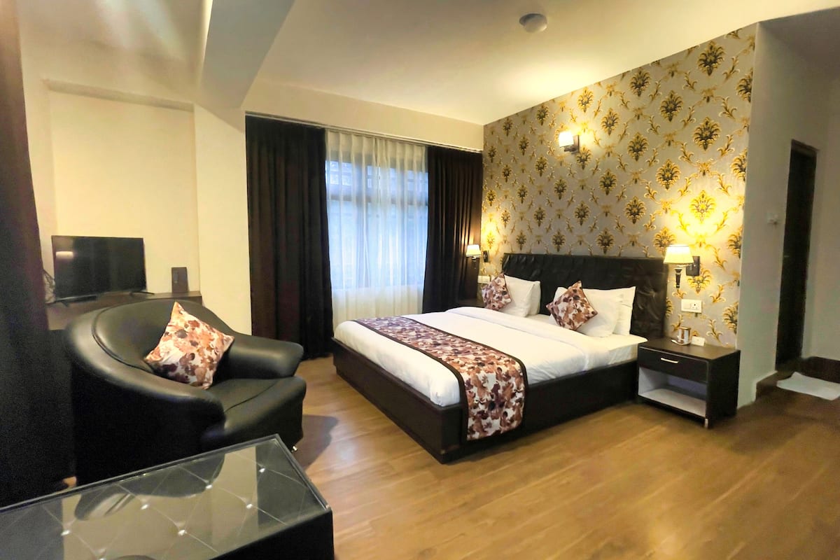 A hotel room with a large bed, a black leather armchair, a glass coffee table, and a nightstand with a lamp.