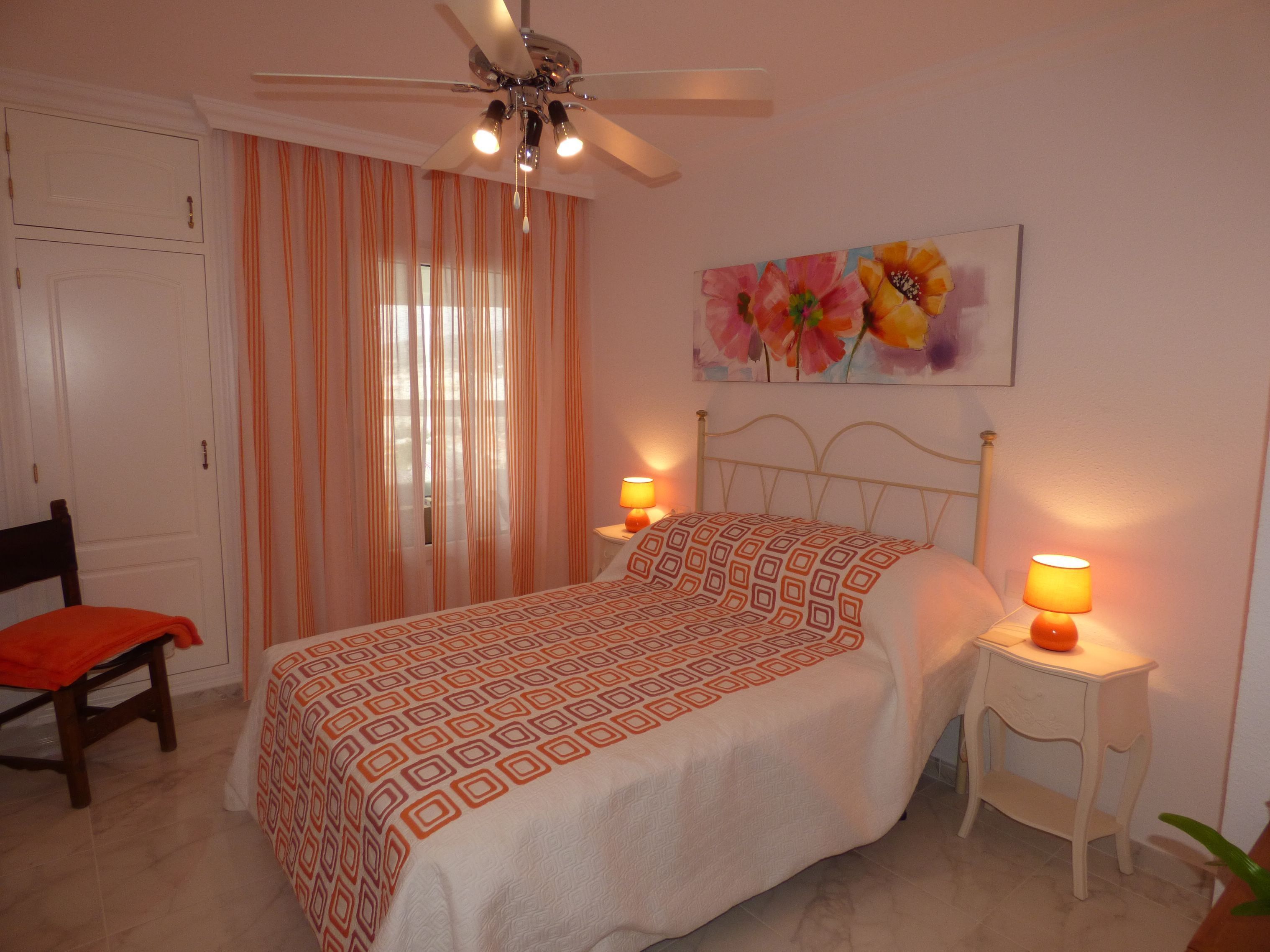 Un dormitorio con cama, dos mesitas de noche con lámparas, ventilador de techo, una silla y ventana con cortinas.