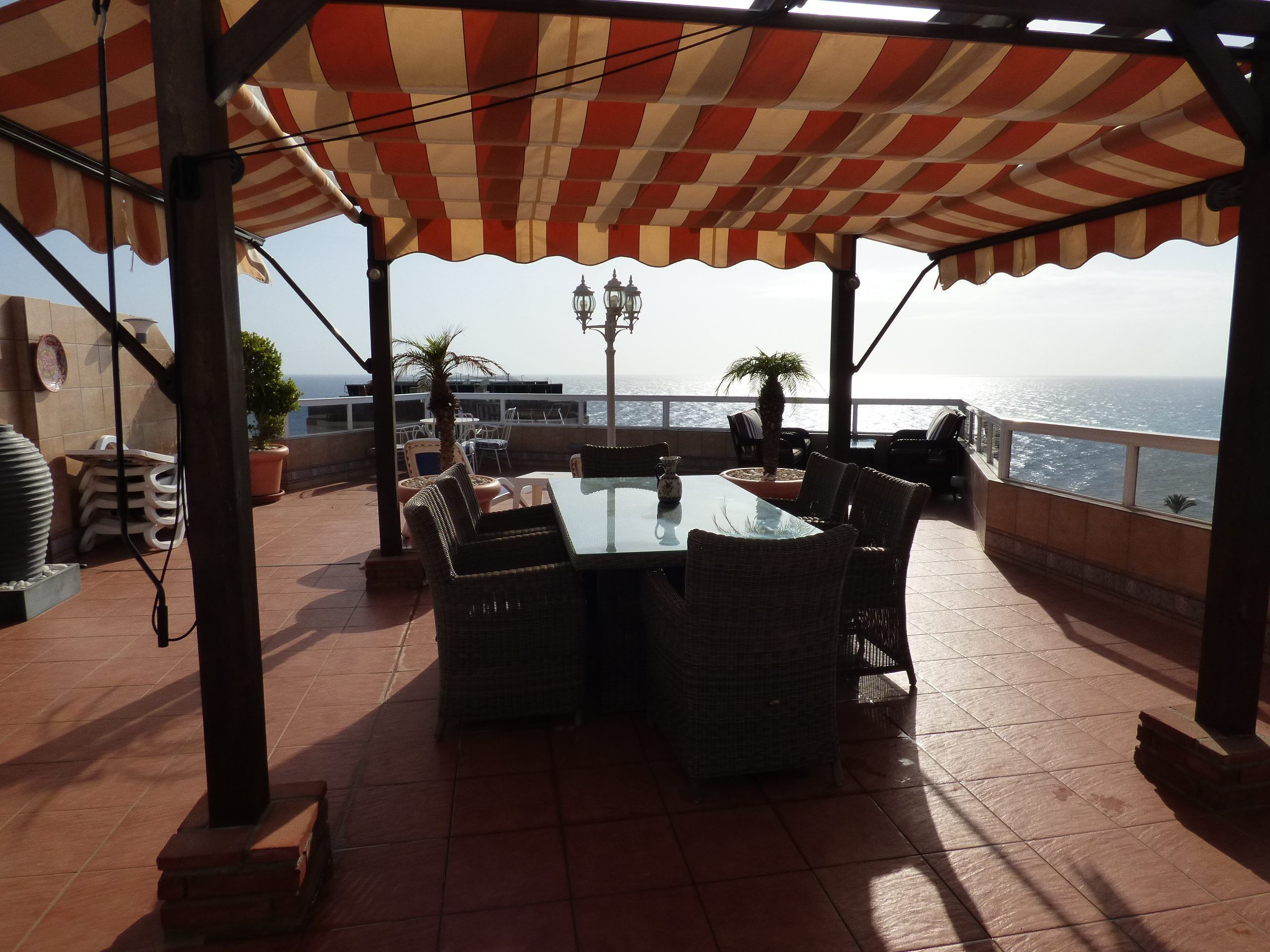 Une terrasse extérieure avec une table et des chaises, un auvent à rayures et une vue sur la mer.