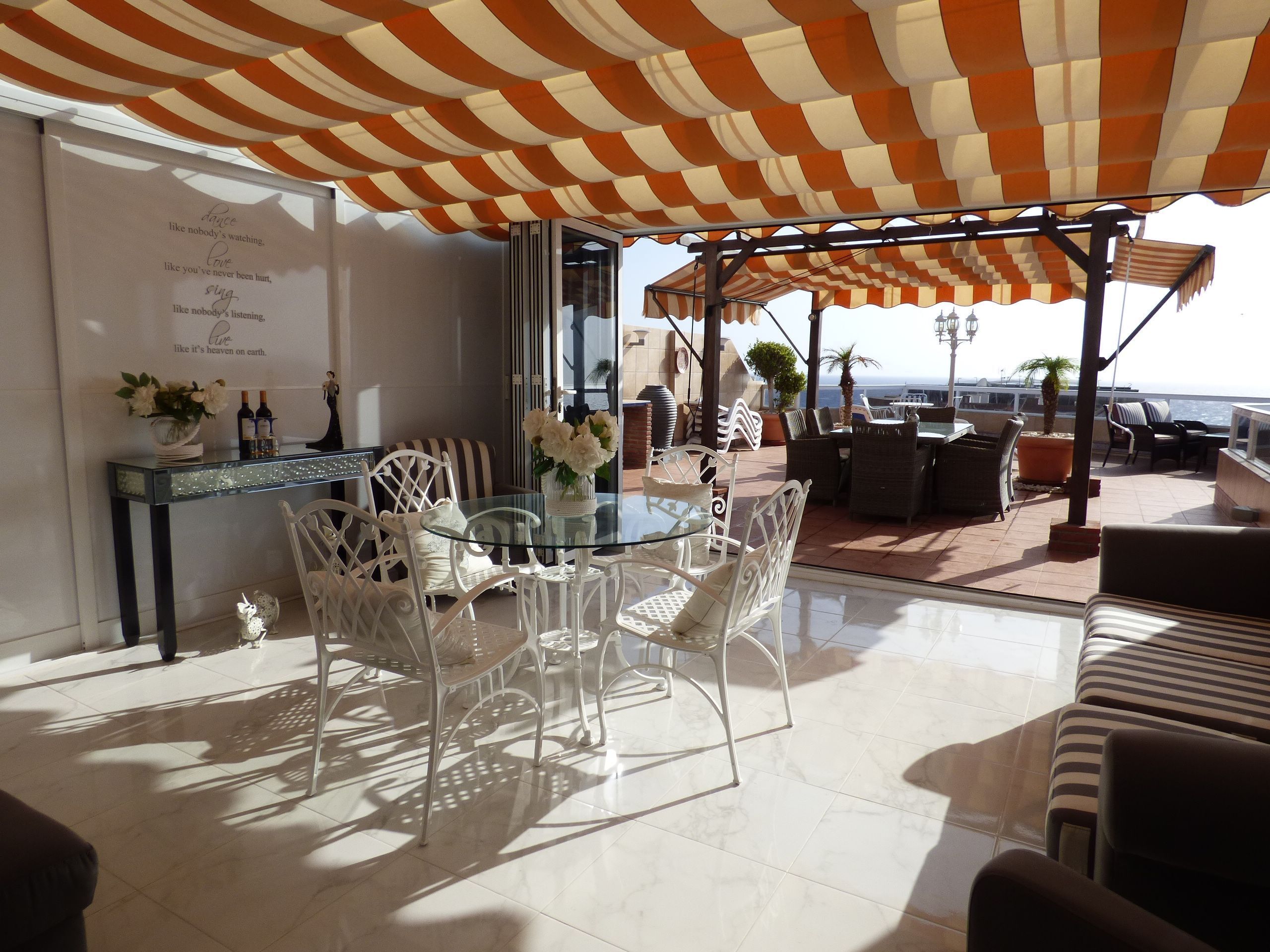 A sunlit terrace with a glass table and white chairs, a striped awning overhead, and a view of the sea.