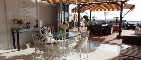 A sunlit terrace with a glass table and white chairs, a striped awning overhead, and a view of the sea.
