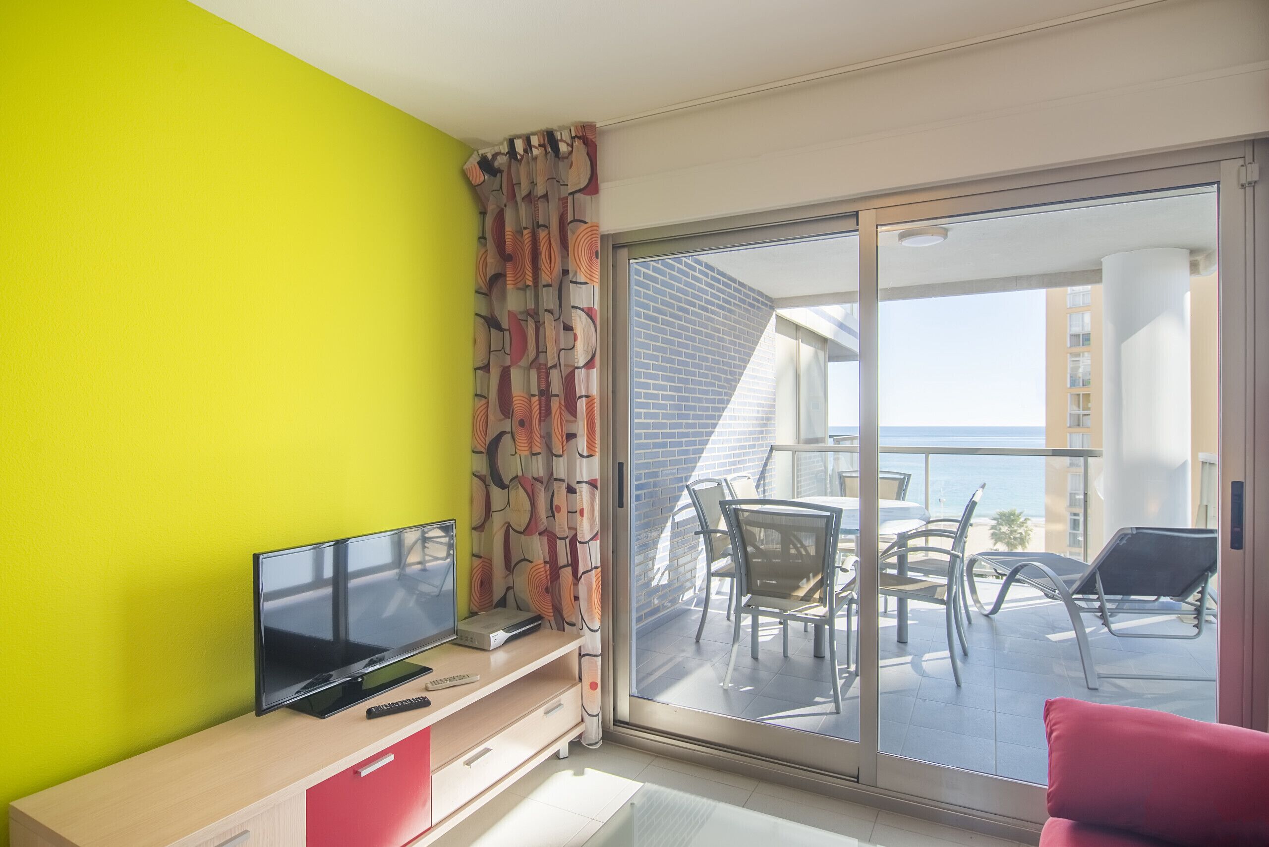 Habitación moderna con una pared de color amarillo brillante, un televisor de pantalla plana, un escritorio pequeño y un puerta corrediza de vidrio que da a un balcón exterior con vistas al mar.