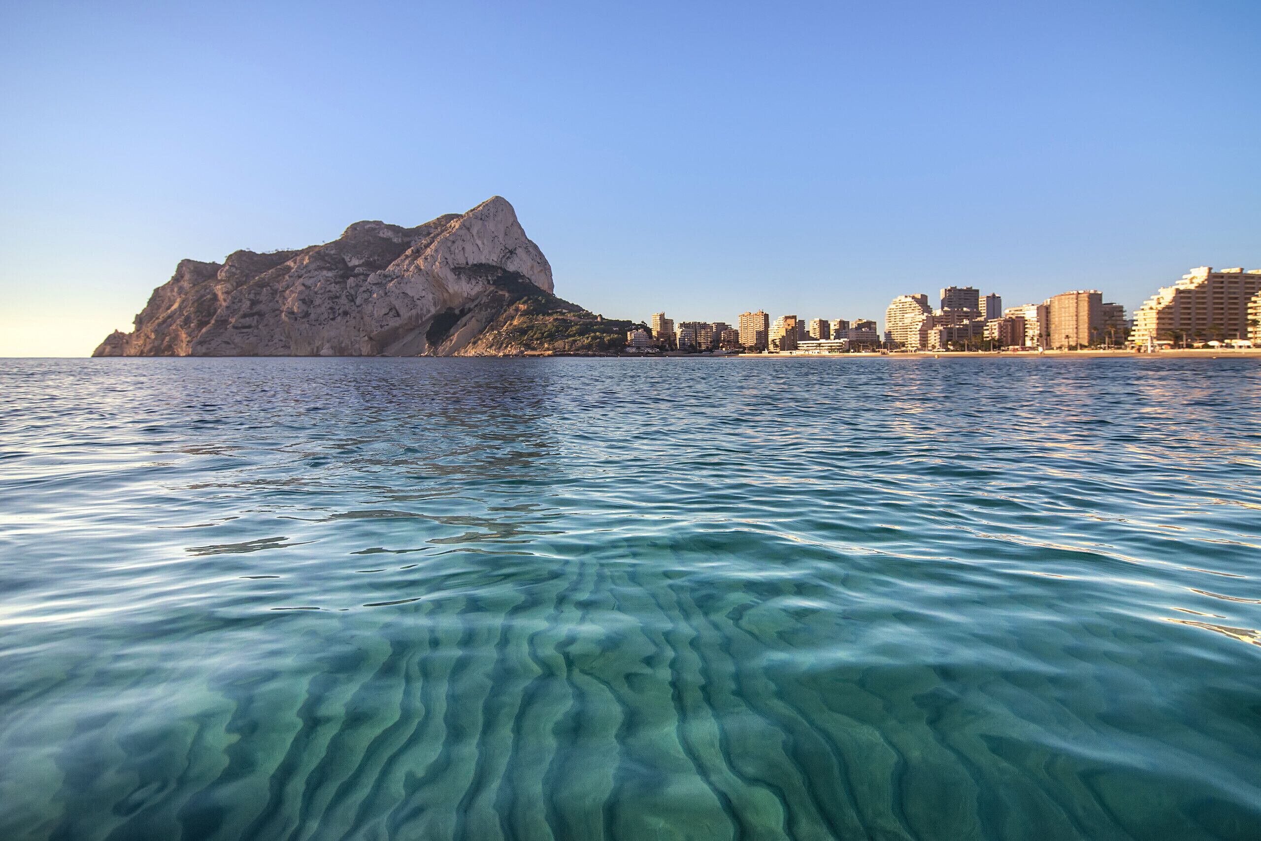 A coastal cityscape with clear waters and a prominent rocky hill.