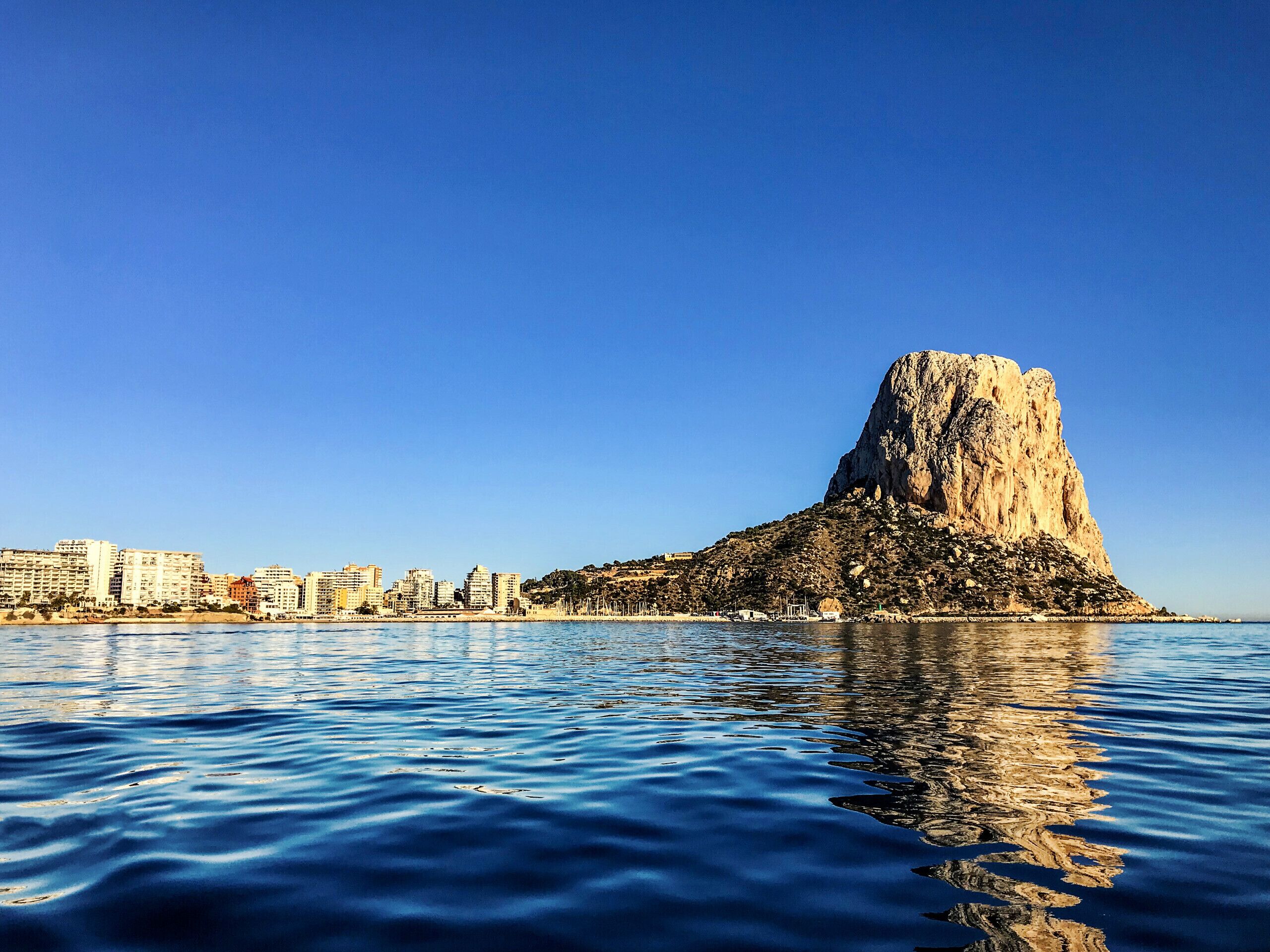 Un paisaje urbano costero con una colina rocosa destacada y un mar tranquilo.