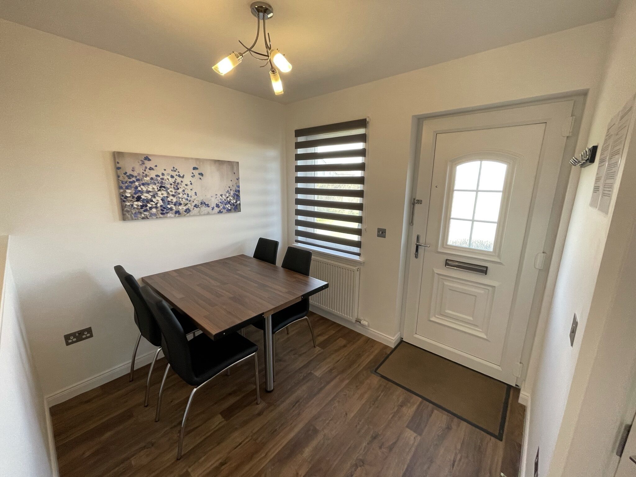 Comedor con mesa de madera y sillas negras, lámpara de techo, ventana con persianas y una puerta blanca.