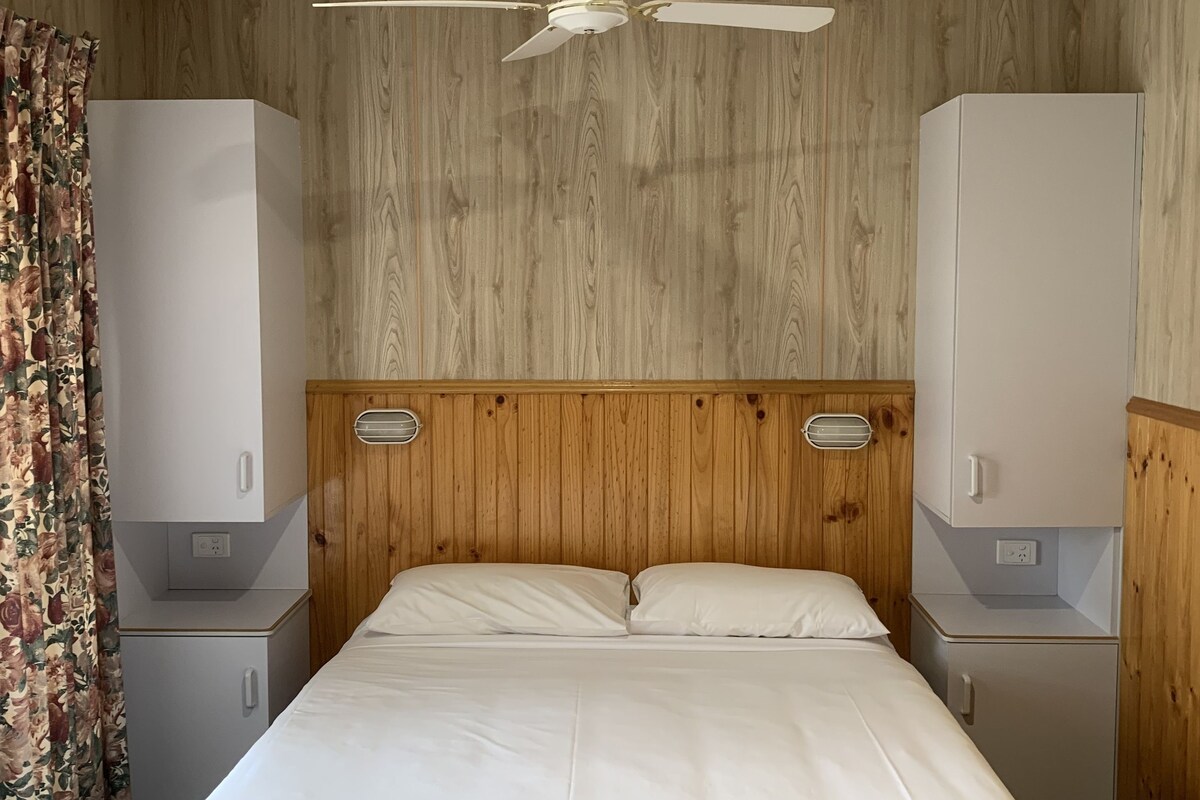 A hotel room with a wooden headboard, a single bed with white bedding, a ceiling fan, and two bedside tables with storage cabinets.