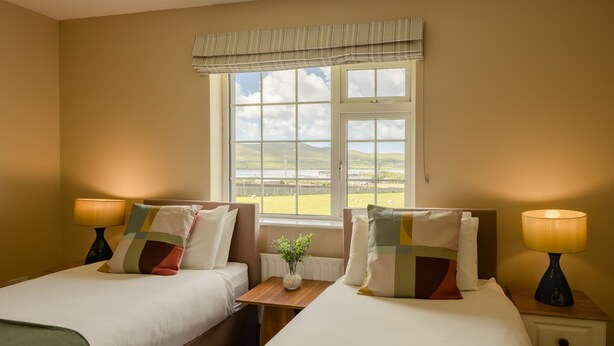 A hotel room with two beds, a window with a view of the countryside, a small table with a vase, and bedside lamps. - Seaview Heights (Dingle)