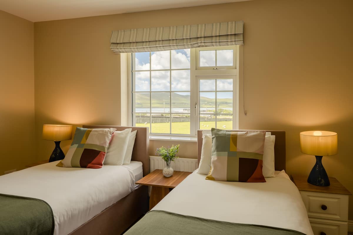 Quarto de hotel com duas camas, janela com vista para o campo, uma mesinha com um vaso e luminárias de cabeceira.