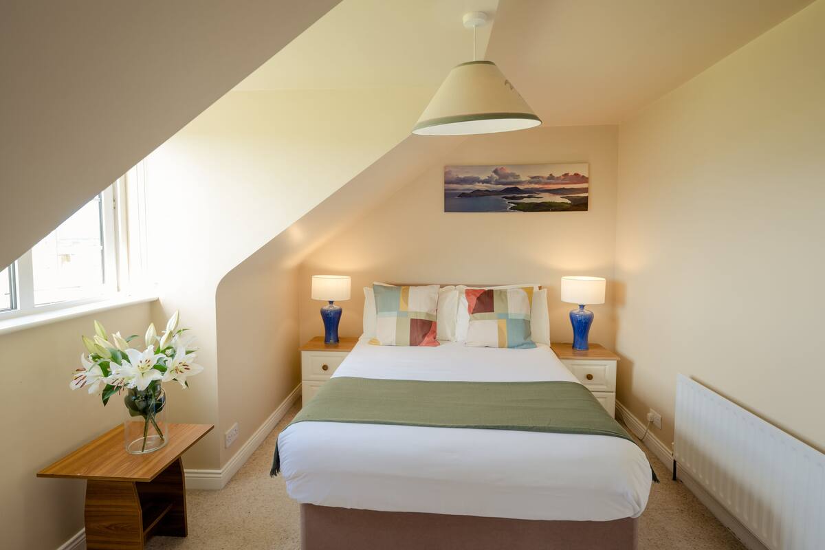 Quarto com cama, mesas de cabeceira, luminárias, um quadro e um vaso com flores.