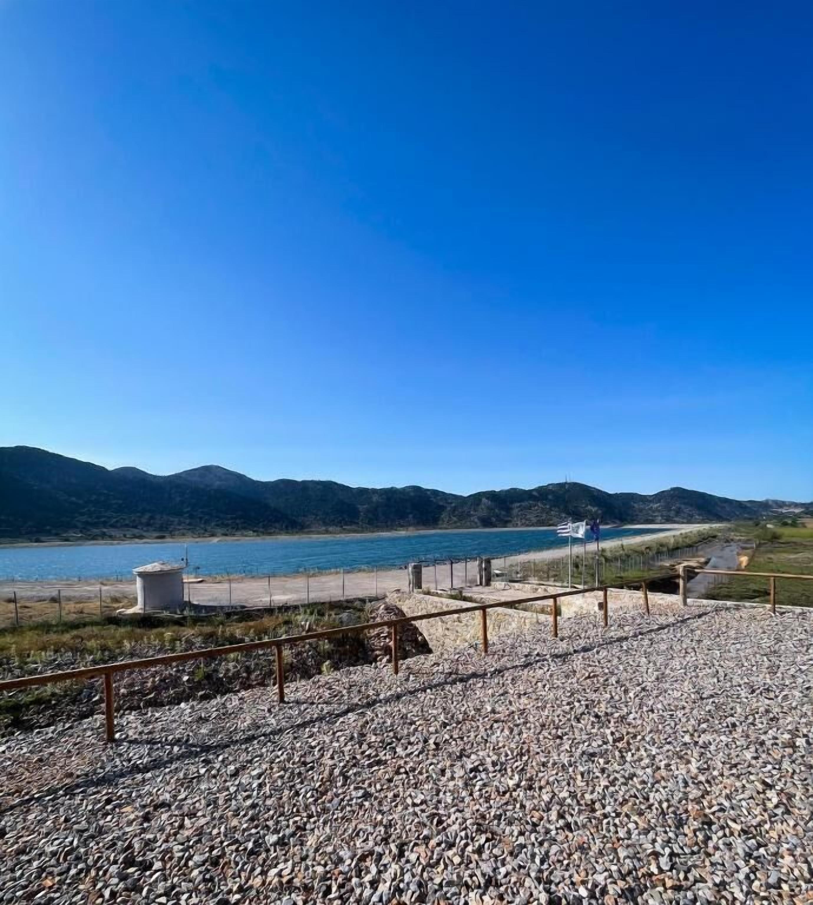 Ein Kiesstrand mit einer Umzäunung und freiem Blick auf das Wasser und ferne Berge.