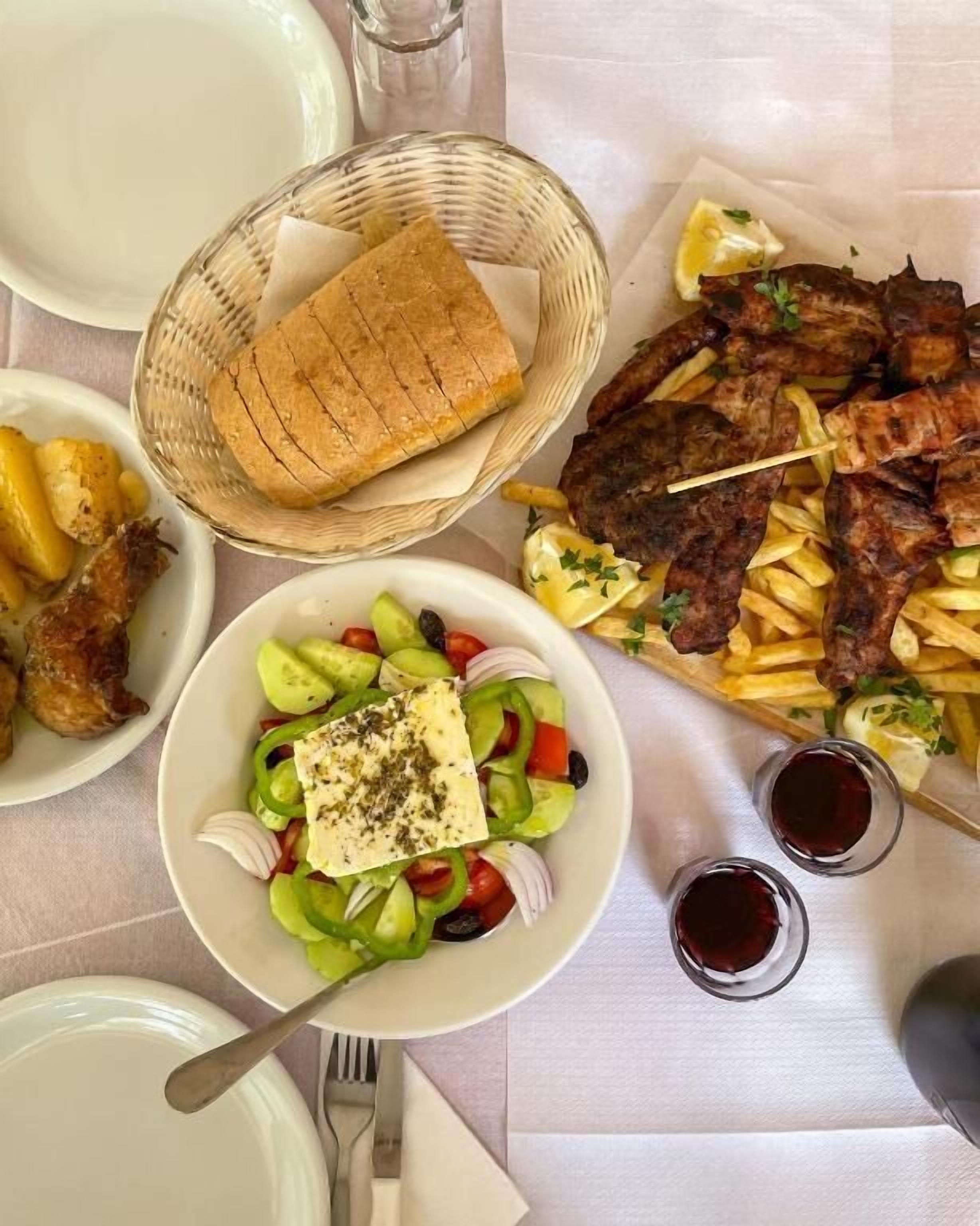 A table set with plates of food, including a salad, grilled meat, and fries, along with a basket of bread and glasses of a dark beverage.
