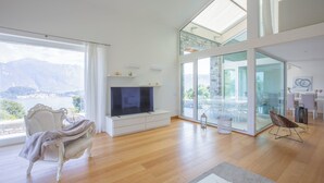 Un salon spacieux doté d’une grande fenêtre, d’un téléviseur à écran plat, d’un canapé blanc et d’une porte en verre donnant sur un balcon.