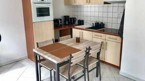 Ett kompakt kök med en inbyggd ugn, mikrovågsugn och kaffebryggare. Köket har ljusa träskåp, en kakelbeklädd vägg bakom arbetsbänken och ett enkelt matbord med stolar.