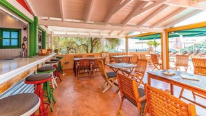 An outdoor cafe with wooden tables and chairs, a bar area with stools, and a view of trees and a building in the background. - Enetiko Resort Hotel (Parga)