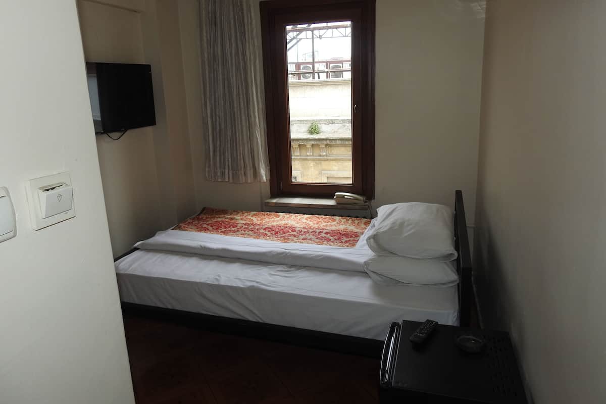 A small, single-bed room with a window, a bedside table, and a wall-mounted TV.