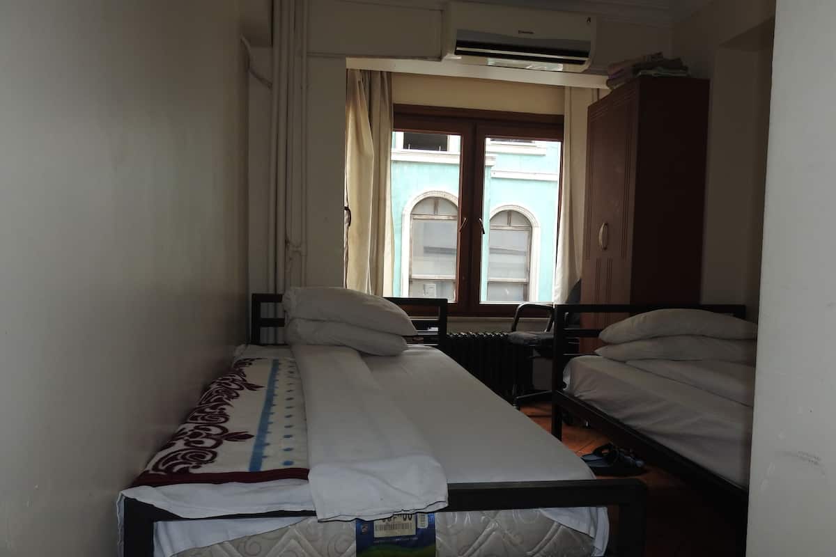 A narrow room with two single beds, a radiator, a window with curtains, and an air conditioning unit.