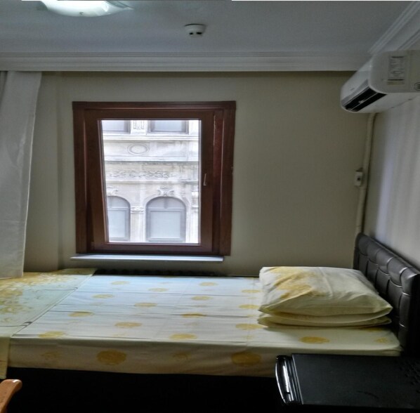 Una habitación pequeña con una cama individual, un ventanal con vista a un edificio y un equipo de aire acondicionado.