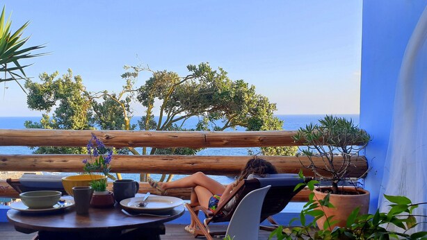 Eine Holzterrasse mit Tisch und Stühlen, mit Blick auf ein Gewässer, im Hintergrund Bäume.
