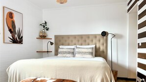 Ein Schlafzimmer mit einem großen Bett, einer hölzernen Bank, einem an der Wand befestigten Regal mit einer Pflanze und einer Pendelleuchte.