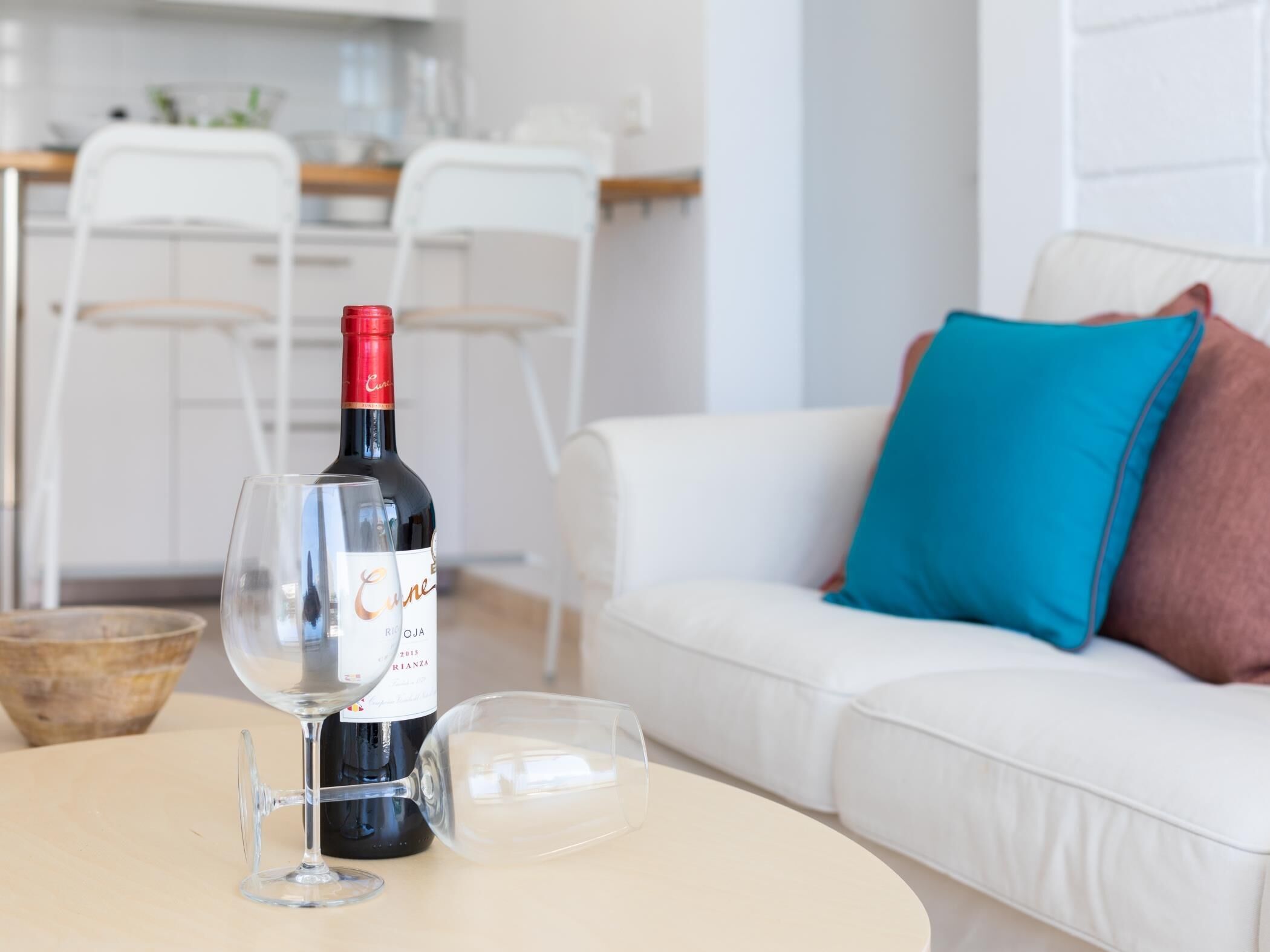 A living room with a white sofa, a blue pillow, a wine bottle, and two wine glasses on a round table.