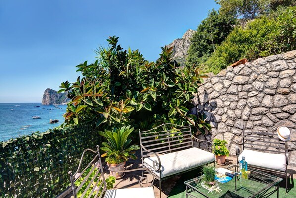 Enjoy the serene sea view from this outdoor seating area, featuring a bench and table amidst lush greenery.