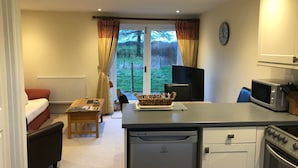 A compact kitchen with a dishwasher, microwave, and stove. There's a dining area with a table and chairs, a sofa, and a window with curtains. - Orchard Cottage. 2-bedroomed cottage. Short breaks welcome. Free Wi-Fi (Hereford)