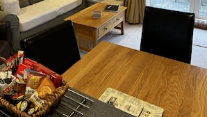 A cozy living room with a sofa, a coffee table, and a dining table. There is a window with curtains, a radiator, and a painting on the wall. - Orchard Cottage. 2-bedroomed cottage. Short breaks welcome. Free Wi-Fi (Hereford)