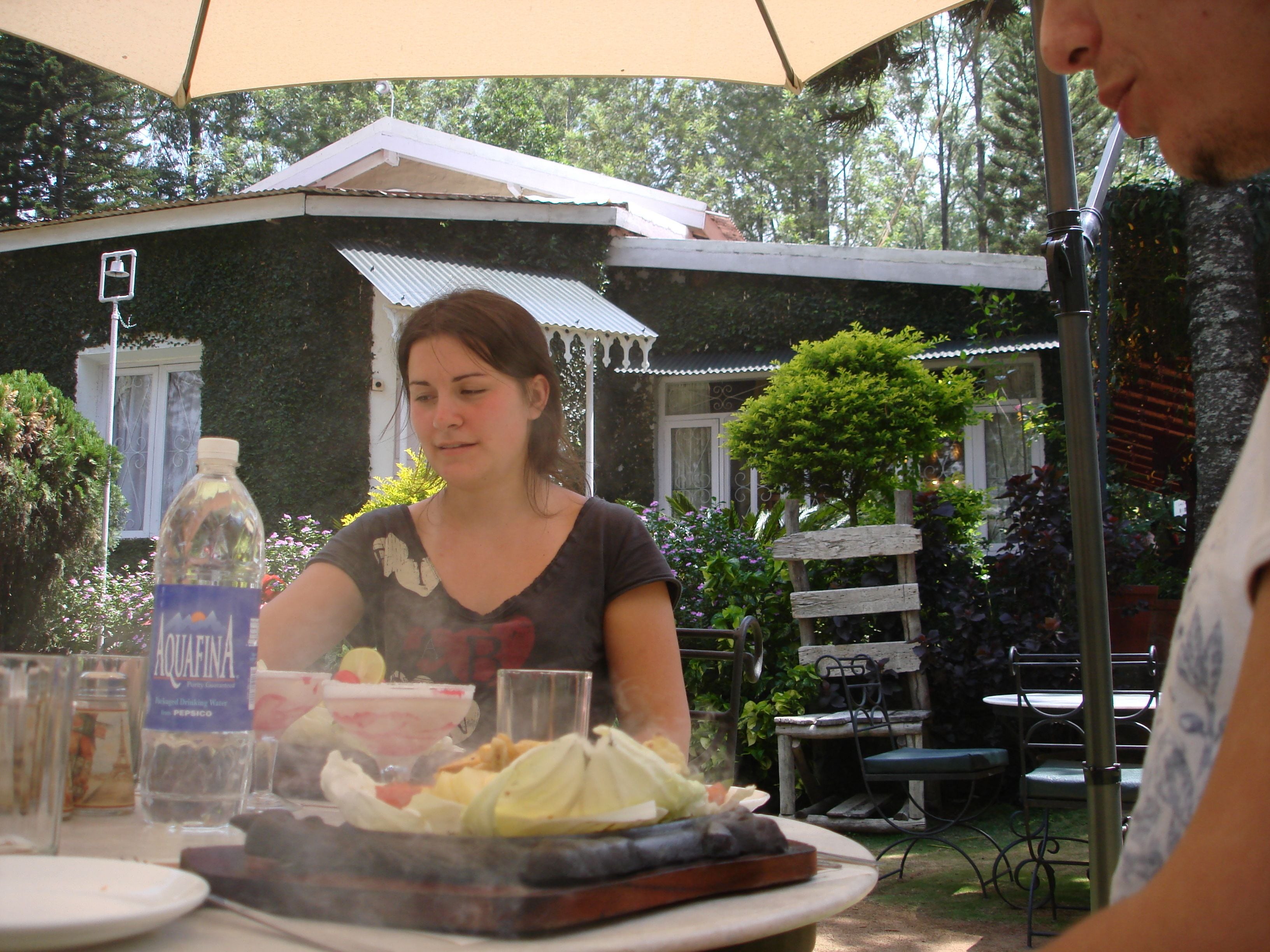 A person seated at an outdoor table with a grill, surrounded by greenery and a white building with a window.