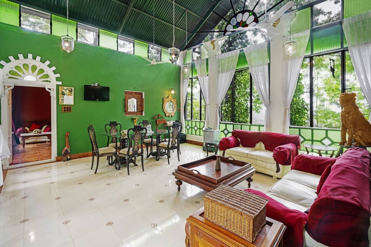 Una habitación con pared verde, zona de comedor con sillas y mesa, un sofá y un ventanal con vistas a árboles.