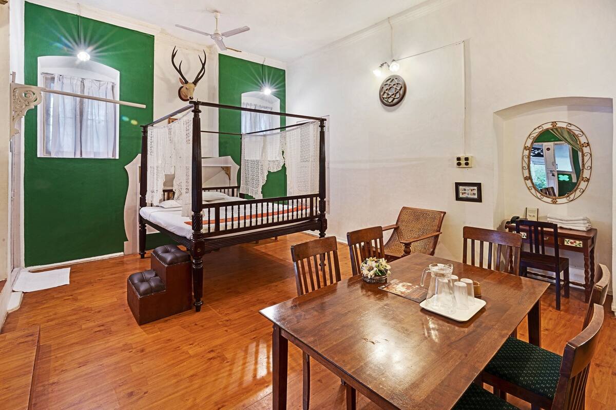 Una habitación con una cama con dosel, una mesa de comedor con sillas, una mesita con una jarrón y vasos, y un espejo en la pared.