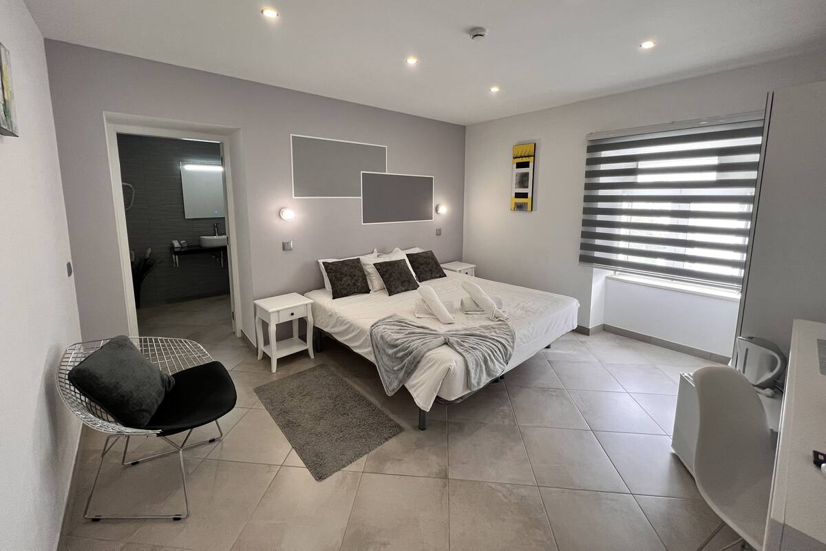 A modern bedroom with a large bed, bedside tables, a chair, a bathroom visible through an open door, and a window with blinds.