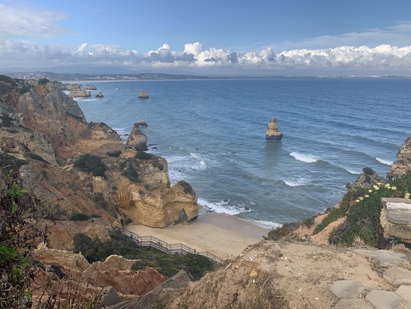 A coastal landscape with rocky cliffs, a sandy beach, and a clear blue sea.