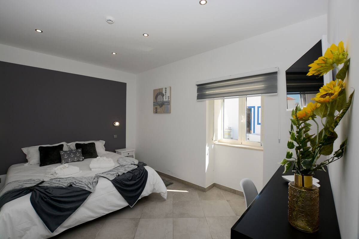 A modern bedroom with a large bed, a bedside table, a window with blinds, and a vase with flowers.