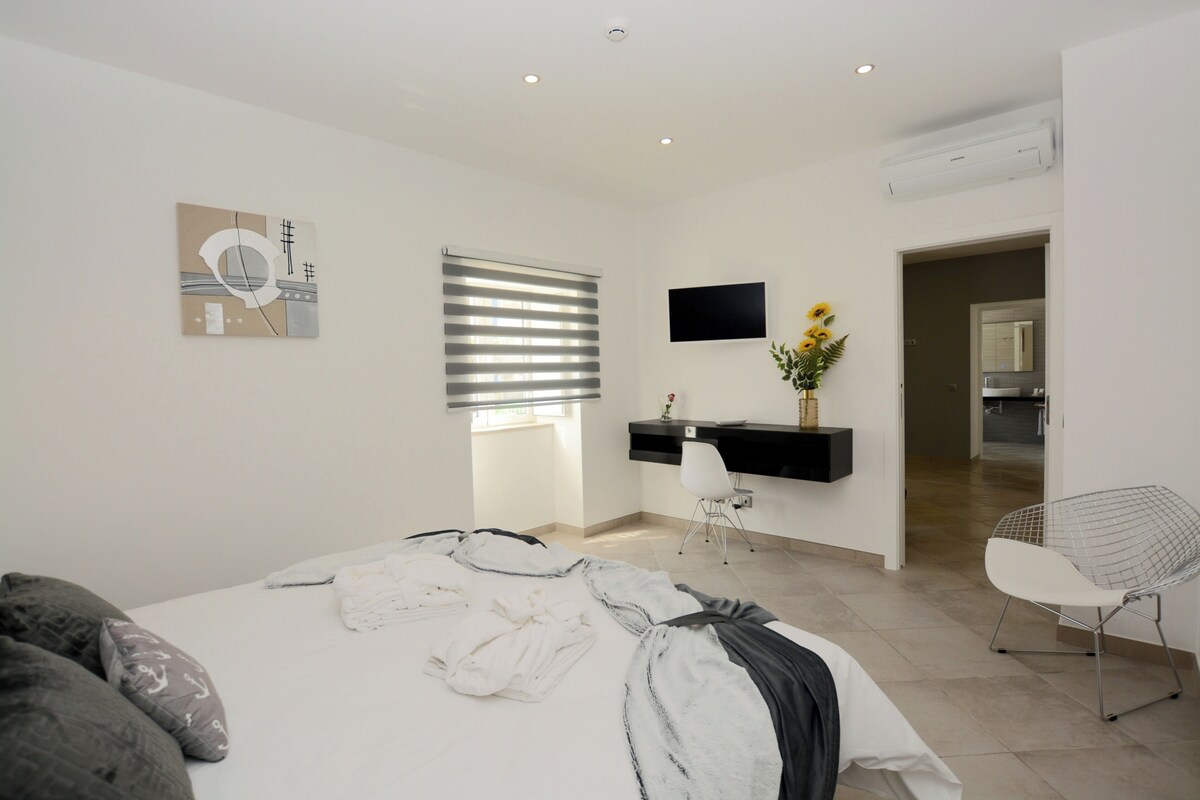 Een moderne slaapkamer met een groot bed, een bureau en een stoel. Er is een televisie aan de muur bevestigd en een raam met zonwering.