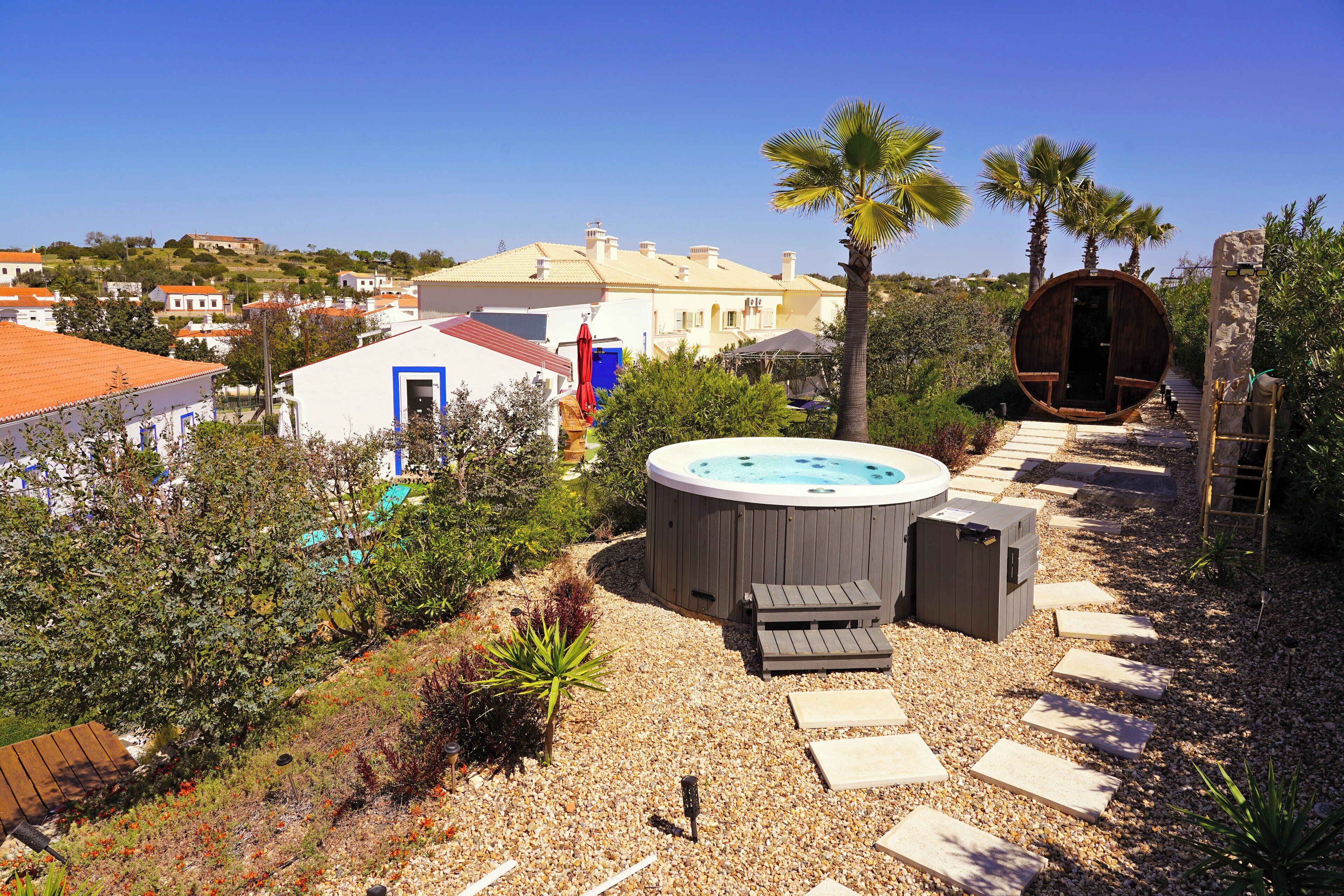 A hot tub with seating, surrounded by a pebble path, palm trees, and a view of houses in the background.