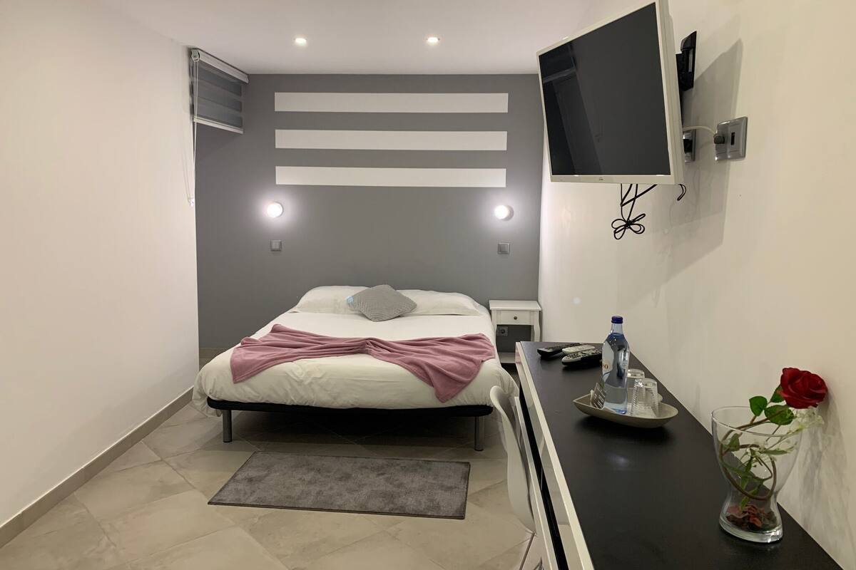 A modern hotel room with a bed, bedside table, TV, and a vase with a rose.
