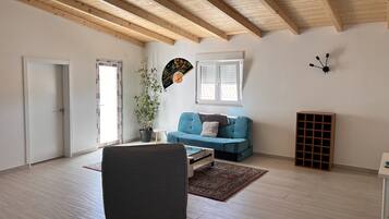 A spacious living room with a high ceiling, wooden beams, a blue sofa, a grey armchair, a small table, and a potted plant.
